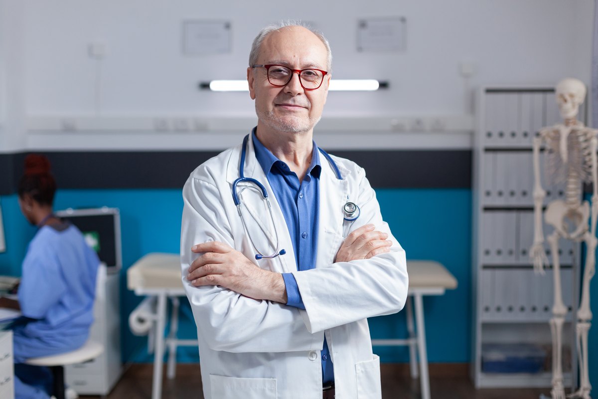 Médico con bata blanca y estetoscopio en una consulta, de pie y mirando a camara