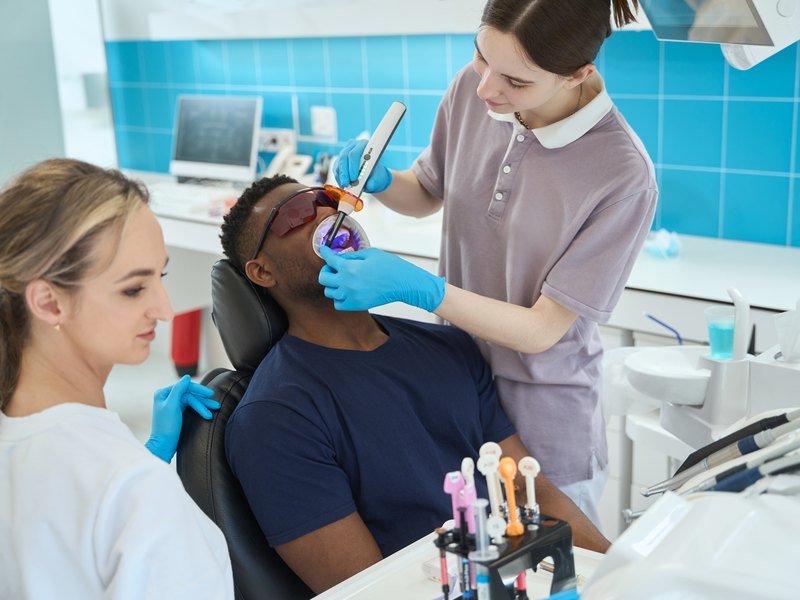 Odontologa y auxiliar tratando a un paciente en el sillon dental con lampara de fotopolimerización